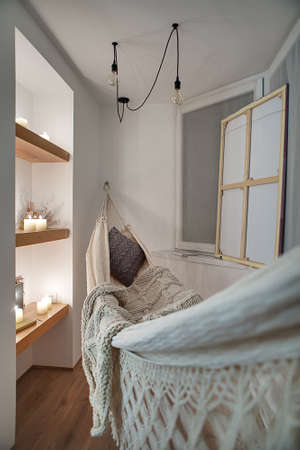 Small room in a modern style with white walls and a parquet on the floor. There are wooden shelves with burning candles and decorations, hammock with a pillow and a plaid, window, canvas and lamps.の写真素材