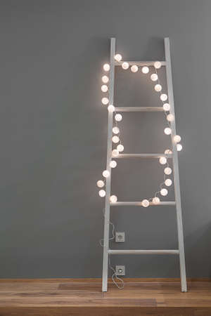 White ladder with glowing sphere lamps in a form of the garland on the gray wall background. There is a parquet on the floor and power sockets on the wall. Closeup. Vertical.の写真素材