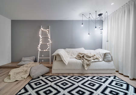 Room in a modern style with white and gray walls and a parquet with a carpet on the floor. There is a sofa, plaids, pillows, wooden ladder with sphere glowing lamps, hanging lamps and decorative owls.の写真素材