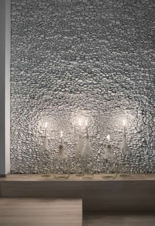 Burning candles in the bottles on the wooden rack on the background of the wall decorated with pebbles. On the floor there is a parquet. Closeup. Vertical.の写真素材