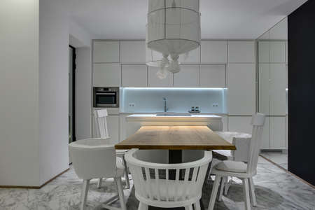 Modern kitchen with white walls and gray tiles with patterns on the floor. There is a wooden table with white chairs, kitchen island with a stove, white lockers, oven, sink with a faucet, mirror.の写真素材