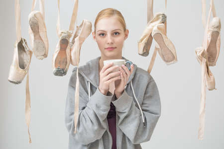 Portrait of a young ballerina with a cup of tea in her hands is among the hanging ballet slippers. Pointe shoes are arranged in random order.の写真素材