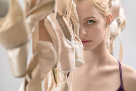Blonde ballerina stands between hanging beige pointe shoes on the light background in the studio. She wears a violet top and looks into the camera. Closeup. Horizontal.の写真素材