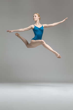 Incredible ballerina posing in the jump on the gray background in the studio. She wears a blue leotard and looks to the side. Her legs and arms stretched to the sides. Vertical.の写真素材