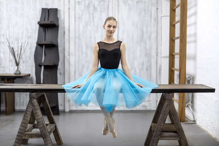 Smiling ballerina sits with crossed legs on the wooden table on a light wall background in a studio. She wears a black leotard with a cyan tutu and pointe shoes. She looks into the camera. Horizontal.の写真素材