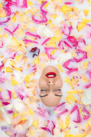 Amazing girl with closed eyes in the bath full of water with milk and colorful flower petals. On the...