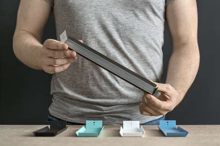 Man in gray T-shirt with a metal dark support in the hands on the dark background in the workshop. On the table under his hands there are other multicolored supports: black, white, mint and blue.の写真素材