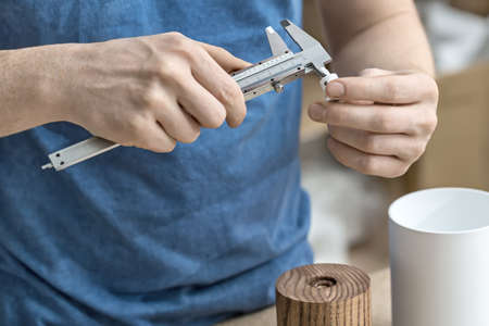 Measuring process in the workshop. Man is using a caliper on the white metal detail. Under his hands there are wooden and metal cylindrical billets. Closeup. Horizontal.の写真素材