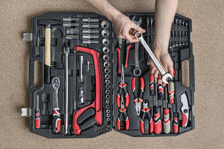 Black toolbox on the light tabletop in the workshop. Man with a tattoo holds a spanner above it. There are black-red pliers and screwdrivers, ratchets, spanners, hammer, saw and different nozzles.の写真素材
