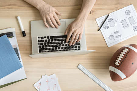 Man is using a laptop on the wooden table in the workshop. Around the laptop there are draughts, drafts, notebook and books, marker and a pencil, ruler, football ball. Top view closeup photo.の写真素材