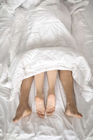 Nice barefoot legs of lovers under the white blanket on the bedの写真素材