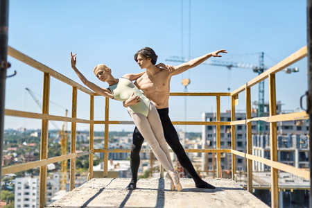 Incredible couple of ballet dancers posing on the concrete balcony of the unfinished building on the cityscape background. Topless guy wears a black dance pants, girl wears a green leotard.の写真素材