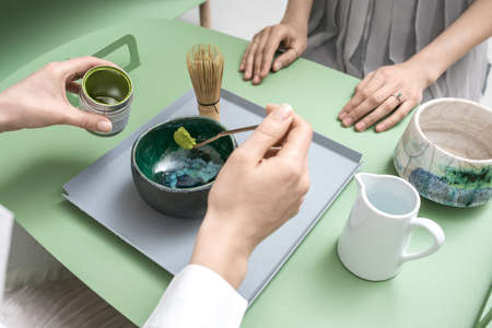 People preparing a chinese matcha green tea on the green metal table indoors. One woman measures tea powder with a wooden stick over the bowl. On the table there is tea whisk and white jug with water.の写真素材