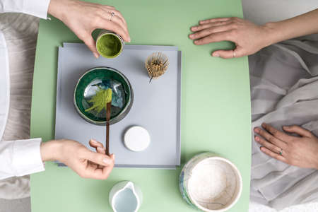 Two girls preparing a chinese matcha green tea on the green metal table indoors. One woman puts tea powder into the colorful bowl. On the table there is a wooden tea whisk and white jug with water.の写真素材
