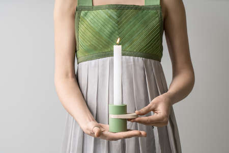 Delightful girl holds a metal-wooden green candlestick with a burning candle in her hands on the light background in the studio. She wears a colorful dress. Closeup. Horizontal.の写真素材