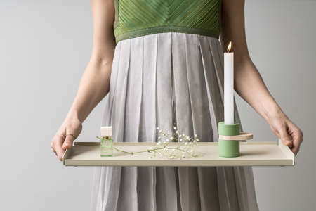 Cute girl in a colorful dress holds a beige metallic support in her hands on light background in the studio. On the support there is green candlestick with burning candle, glass jar, flower's branch.の写真素材
