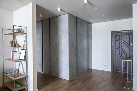 Contemporary hallway with white walls and a parquet on the floor. There are textured gray lockers, dark entrance door, stand with keys and glasses, shelves with books and accessories. Horizontal.の写真素材