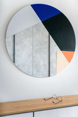 Colorful round mirror on the light wall. Under it there is a wooden stand with glasses. Indoors. Closeup. Vertical.の写真素材