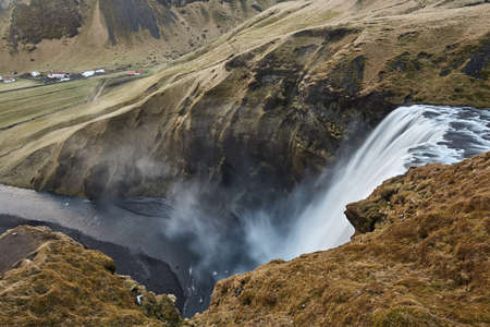 Amazing waterfall falls down from the cliff into the valley in Iceland. Cliff covered by faded grass. River flowing into the valley with small settlement. Horizontal.の写真素材