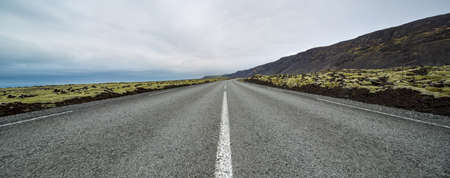 Driveway with orange roadside pillars between the green fields and mountains on the background of the cloudy sky in Iceland. Panoramic. Horizontal.の写真素材
