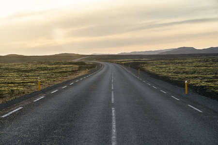 Textured roadway with orange roadside pillars between the green fields on the background of the mountains and cloudy sunset sky in Iceland. Horizontal.の写真素材