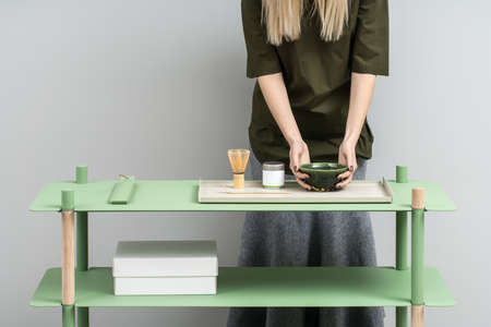 Pretty girl holds an emerald bowl on the green metallic-wooden stand on the gray wall background in the studio. On the stand there is white box, colorful metal supports, jar, tea whisk. Horizontal.の写真素材