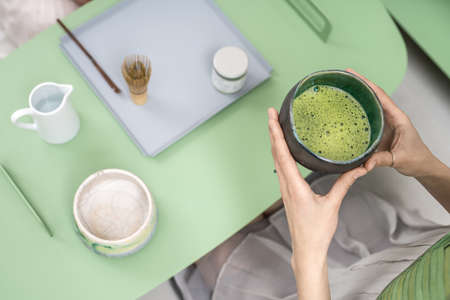 Nice girl holds an emerald bowl with matcha green tea in her hands over the green metallic table indoors. On the table there is a white jug with water, jar, wooden stick and tea whisk. Closeup.の写真素材