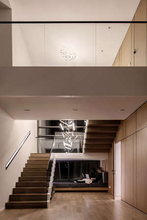 Cool modern interior with light walls and a stair with wooden rungs and a glass railing. There are hanging luminous fancy lamps and a dark sofa with a woman with a laptop, opened door. Vertical.の写真素材