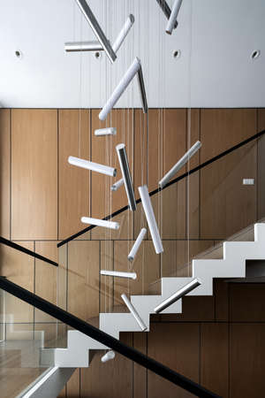Modern interior with light walls and a stair with wooden rungs and glass railing. There are hanging fancy lamps. Vertical.の写真素材