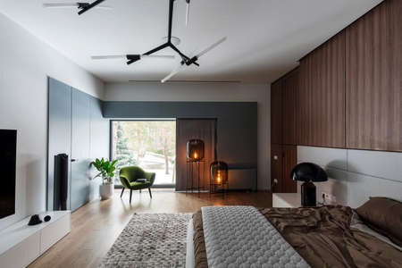 Modern bedroom with white walls and a parquet with a carpet on the floor. There is a bed with a brown linens and a gray plaid, black lamp on a stand, wooden lockers, luminous bulb lamps, armchair.の写真素材