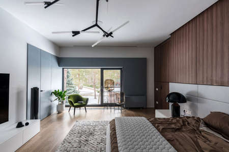 Stylish bedroom with white walls and a parquet with a carpet on the floor. There is a bed with a brown linens and a gray plaid, black lamp on a stand, wooden lockers, bulb lamps, armchair, plant.の写真素材
