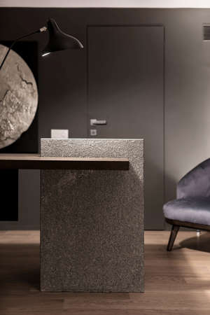Interior in a modern style with gray wall and a parquet on a floor. There is a wooden-stone table with a luminous black lamp, dark armchair, door. Vertical.の写真素材