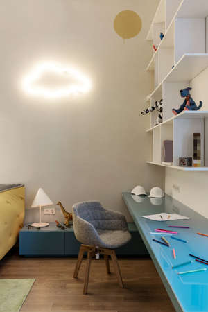 Colorful modern kid's room with white walls and a parquet with a carpet on a floor. There is a blue tabletop with pencils and caps, white shelves with toys and books, chair, luminous fancy cloud lamp.の写真素材