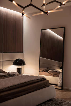 Modern bedroom with white and wooden walls, parquet with a carpet on a floor and luminous fancy lamps. There is a bed with a brown linens and a gray plaid, mirror, night stand. Vertical.の写真素材