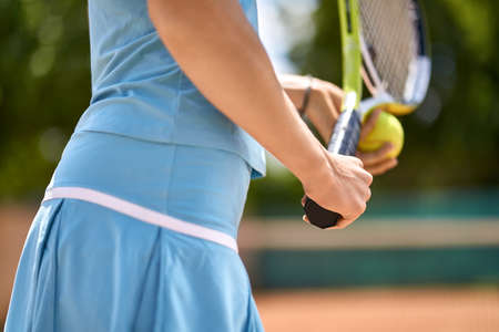 Woman plays tennis on the court outdoors. She holds a racket and a ball. Girl wears a light blue sportswear. Closeup. Horizontal.の写真素材