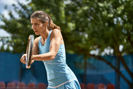 Athletic girl plays tennis on the court outdoors. She prepares to beat off a ball. Woman wears a light blue sportswear. Horizontal.の写真素材