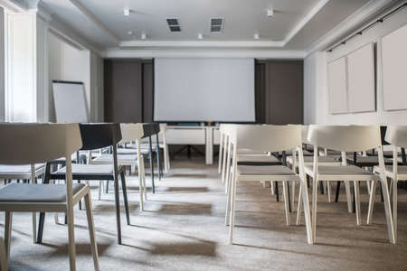 Beautiful conference hall with white walls and a gray floor. There are many light and dark chairs, boards, tables with blue chairs, lamps, window. Horizontal.の写真素材
