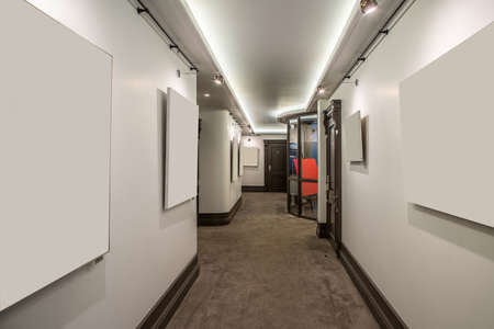 Luminous corridor with light walls and a beige carpet on the floor in the hotel. There are brown doors to the rooms, glass partition, hanging lamps. Horizontal.の写真素材