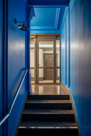 Illuminated interior in the hotel with blue and light walls and a tiled floor. There is a stair with a handrail, glass entrance, brown door with a room number, hanging lamp on the wall. Vertical.の写真素材