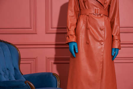 Girl is posing next to a blue armchair on the coral wall background in a studio. She wears a leather orange trench and light blue gloves. Closeup horizontal photo.の写真素材