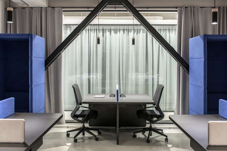 Nice illuminated office in a loft style with gray walls. There are black tables with partitions, dark chairs, two fancy blue armchairs, glass doors with curtains, hanging lamps. Horizontal.の写真素材