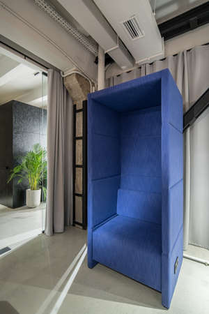 Zone for relax in an illuminated office in a loft style. There is a fancy blue armchair, glass door with curtains, concrete column, green plant in a big pot. Vertical.の写真素材