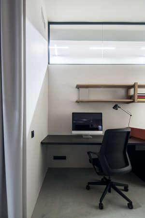 Room with luminous lamps in a office in a loft style with gray walls. There is a black table with a peach partition, dark chair, computer, wooden shelves with books, wide mirror on the wall.の写真素材