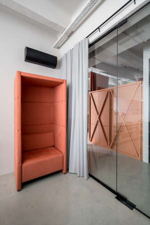 Relax zone with a fancy peach armchair in an office in a loft style with gray walls. There is a glass door with curtain and a black conditioner on the wall. Vertical.の写真素材