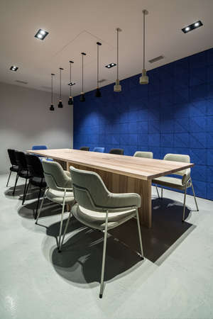 Conference room with luminous lamps in an office with gray and textured blue walls. There are wide wooden tables with multicolor chairs, hanging dark and light lamps. Vertical.の写真素材