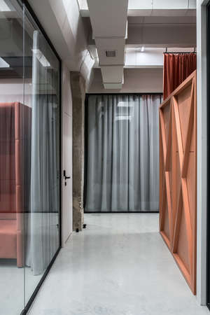 Hall in a luminous office in a loft style with gray walls and a glossy floor. There are glass partitions with doors and curtains, orange partition, concrete column. Vertical.の写真素材