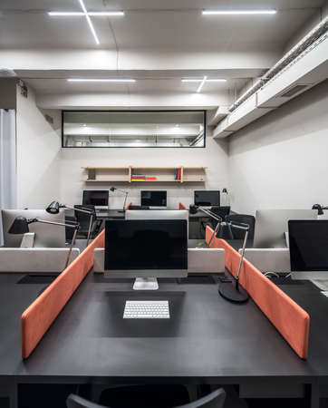 Nice luminous interior in an office in a loft style with light walls. There are black tables with peach partitions, dark chairs, computers, lamps, wooden shelves with books, glass partition on a wall.の写真素材