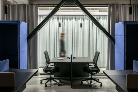 Stylish illuminated office in a loft style with gray walls. There are black tables with partitions, dark chairs, two fancy blue armchairs, glass doors with curtains, hanging lamps. Horizontal.の写真素材