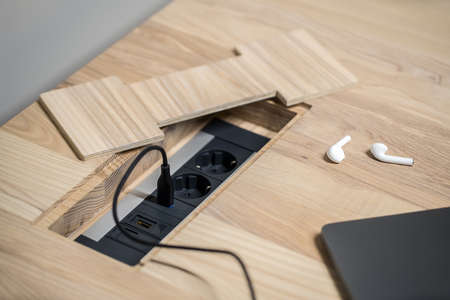 Black power sockets built-in the light wooden table on the gray wall background. On the table there is a laptop with a power cable and white wireless earphones. Closeup. Horizontal.の写真素材