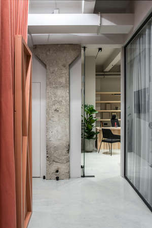 Rooms in a luminous office in a loft style with gray walls and a concrete column. There is glass partition with curtains, orange partition, doors, table with black chairs, green plant, shelves.の写真素材